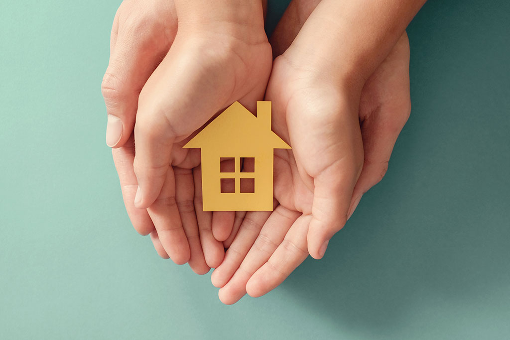 Hands holding yellow paper house on blue background, family home